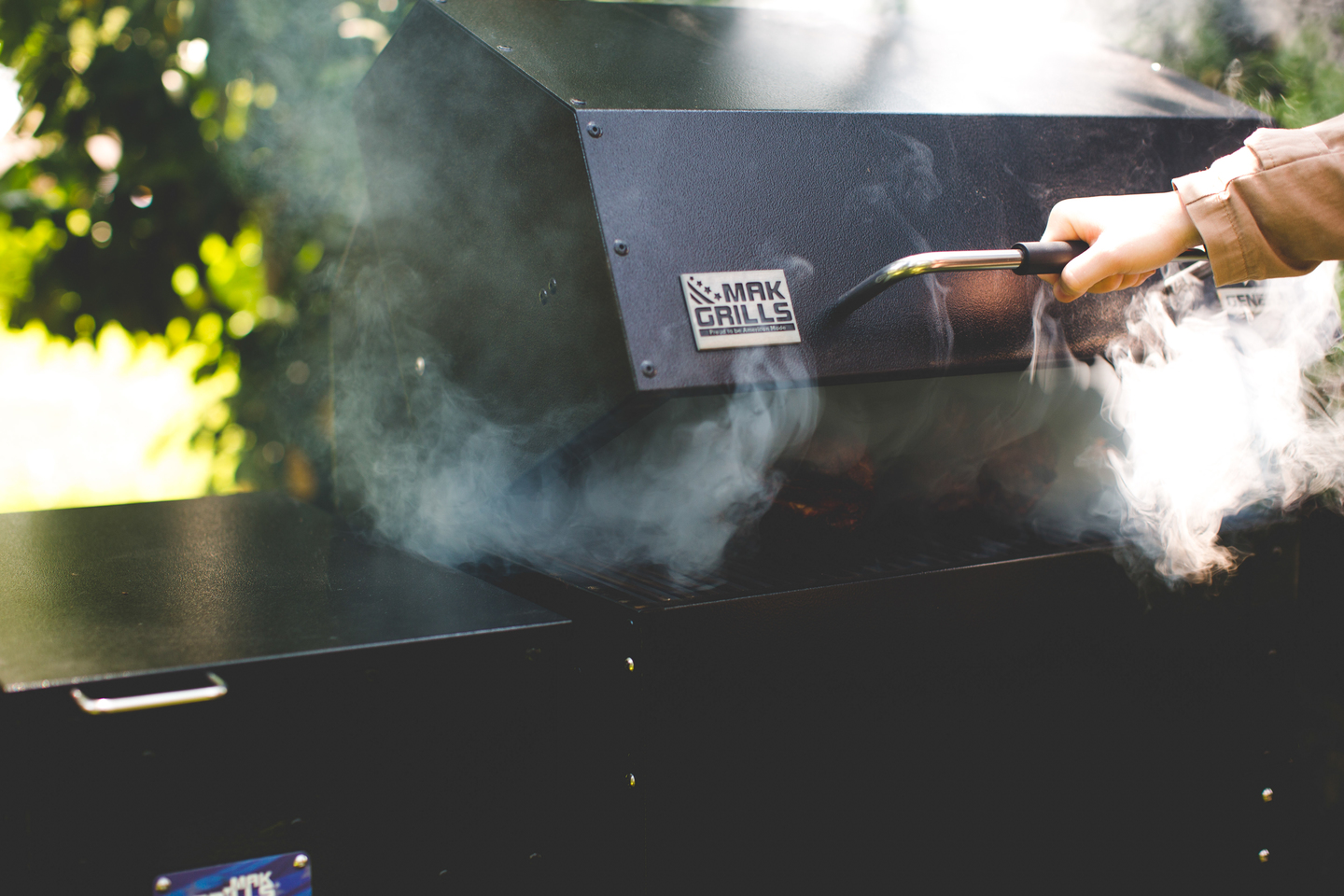 Person lifting the lid of a black MAK Grill with smoke flowing out