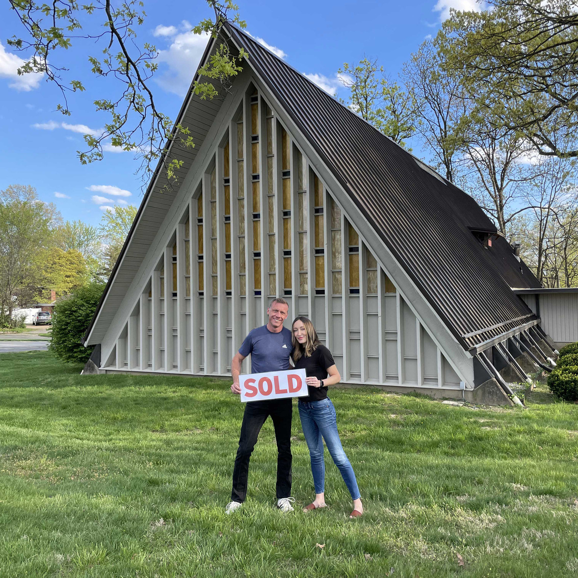 Brenna and her husband holding a "SOLD" sign in front of a church that they bought