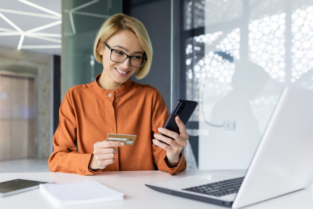 Young businesswoman inside the office at the workplace, holding a phone and a bank credit card in hands, female employee sitting at the table makes online purchases and money transfers.