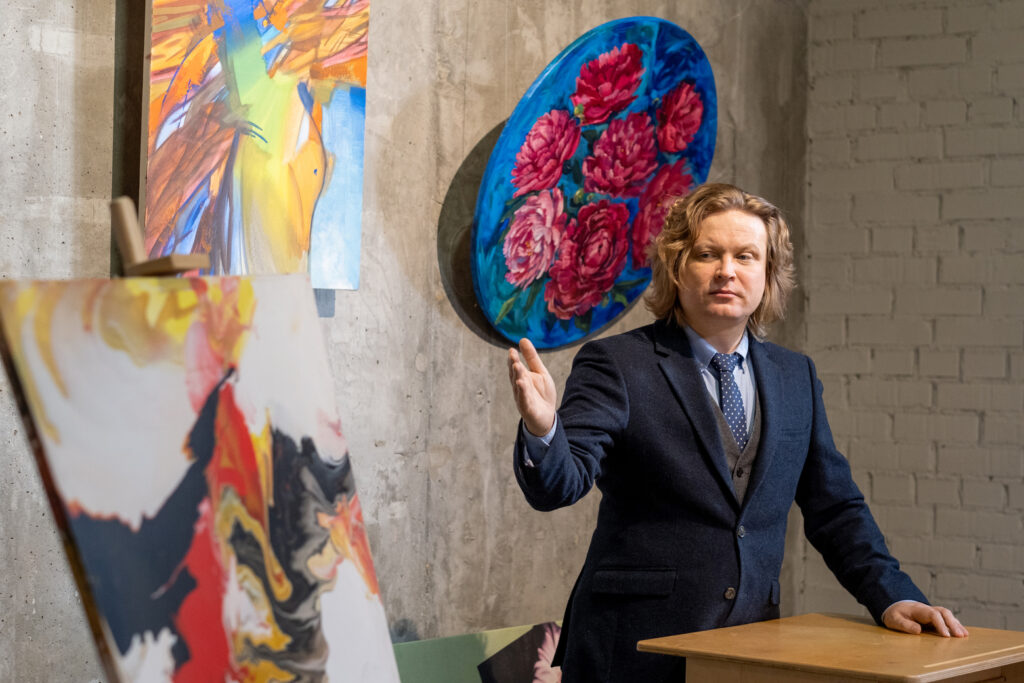 Confident auctioneer in elegant suit standing by tribune while presenting artworks to audience at sale auction